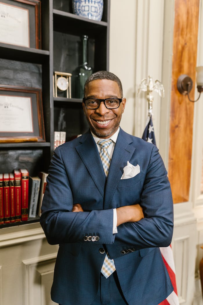 Smiling man with glasses standing confidently in an office with arms crossed.